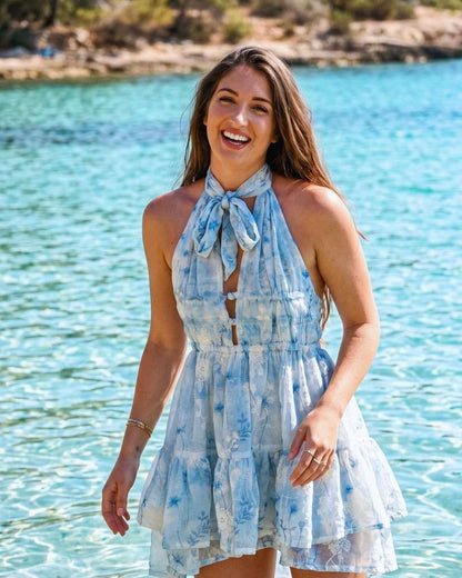 Mujer sonriendo vestida con vestido corto artesanal acuarela azul en una playa y fondo de agua cristalina.
